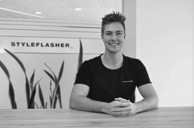 Schwarzweiß-Porträt: styleflasher_-Mitarbeiter Fabio Knoll lächelnd am Tisch; Firmenlogo und Pflanzen im Hintergrund.